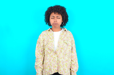 Obraz na płótnie Canvas Dismal gloomy rejected young girl with afro hairstyle wearing floral shirt over blue background has problems and difficulties, curves lower lip and closes eyes in despair, being in depression