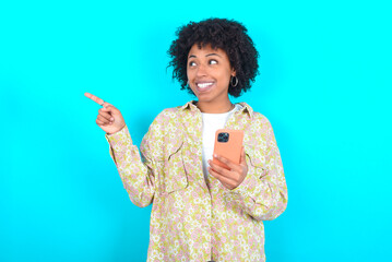 Wow!! excited young girl with afro hairstyle wearing floral shirt over blue background showing mobile phone with open hand gesture