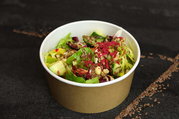Vegetarian salad with cucumbers, lettuce, walnuts and flax seeds in a round takeaway paper container on dark background.Healthy food, restaurant dish delivery. Top view. Free space for text