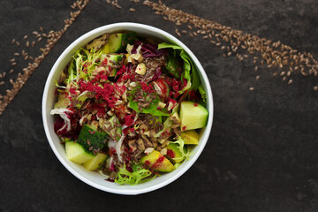 Vegetarian salad with cucumbers, lettuce, walnuts and flax seeds in a round takeaway paper container on dark background.Healthy food, restaurant dish delivery. Top view. Free space for text
