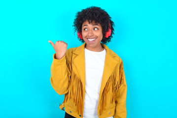 Fototapeta na wymiar Young woman with afro hairstyle wearing yellow fringe jacket over blue background listens audio track via wireless headphones points thumb away advertises copy space