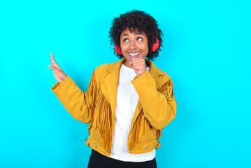 Fototapeta na wymiar Happy Young woman with afro hairstyle wearing yellow fringe jacket over blue background sings favourite song keeps hand near mouth as if microphone wears wireless headphones, listens music
