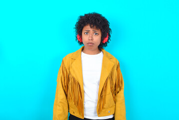 Obraz premium Serious displeased Young woman with afro hairstyle wearing yellow fringe jacket over blue background looks puzzled at camera being angry wears stereo headphones listens music while walking at street