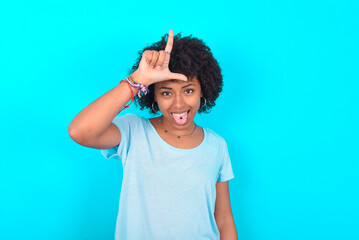 Funny Young woman with afro hairstyle wearing blue T-shirt over blue background makes loser gesture...