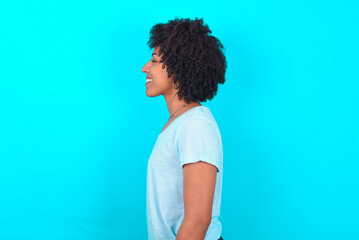 Profile portrait of nice Young woman with afro hairstyle wearing blue T-shirt over blue background look empty space toothy smile © Roquillo