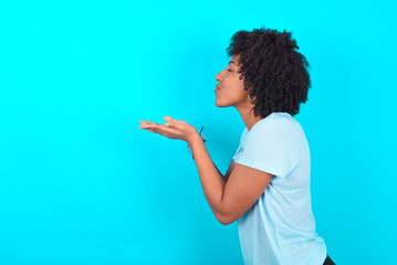 Profile side view view portrait of attractive Young woman with afro hairstyle wearing blue T-shirt over blue background sending air kiss