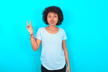 Young woman with afro hairstyle wearing blue T-shirt over blue background makes peace gesture keeps...