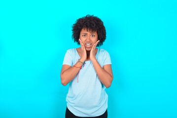 Upset Young woman with afro hairstyle wearing blue T-shirt over blue background touching face with two hands