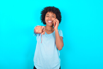 Positive Young woman with afro hairstyle wearing blue T-shirt over blue background indicates directly at camera has telephone conversation smiles broadly enjoys talking long hours. You join me