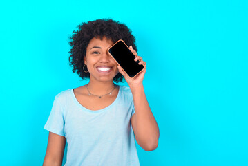 Young woman with afro hairstyle wearing blue T-shirt over blue background holding modern smartphone covering one eye while smiling