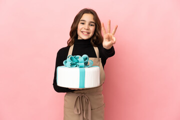 Little Pastry chef holding a big cake isolated on pink background happy and counting three with fingers