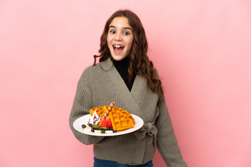 Little girl holding waffles isolated on pink background with surprise facial expression