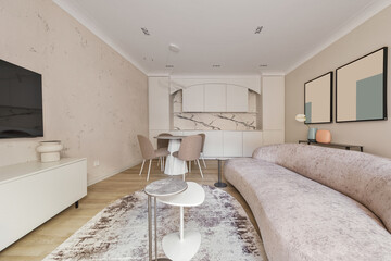 Modern living room interior in pastel colours with round dining table, beige chairs, creamy white kitchen furniture. Trendy minimalistic interior concept