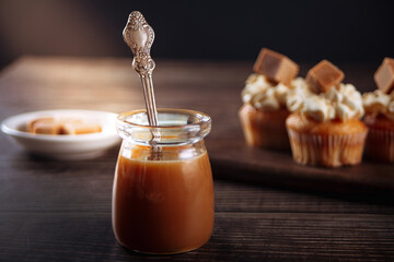 Yummy salted caramel with spoon from glass jar at wooden table and cupcakes on the background.