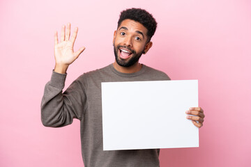 Young Brazilian man isolated on pink background holding an empty placard and saluting