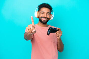 Young brazilian man playing with a video game controller isolated on blue background showing and lifting a finger