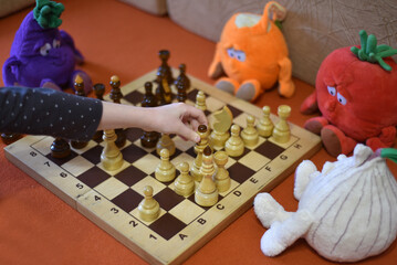 A child's hand rearranges a chess piece on a chessboard. The game is watched by toys sitting nearby on the sofa. 