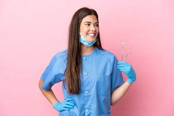 Dentist woman holding tools isolated on pink background posing with arms at hip and smiling