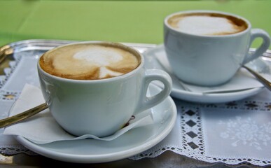 Two cups of cappuccino on a metal tray with a white lace cloth outdoors in summer in Italy.