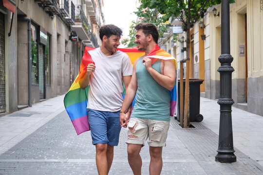 Two young men walking wrapped in a LGBT flag, looking each other and smiling in the street. Gay couple.