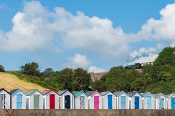 Fototapeta na wymiar Colourful beach houses. Row of multi-coloured beach huts with steam train on stone viaduct against blue sky.