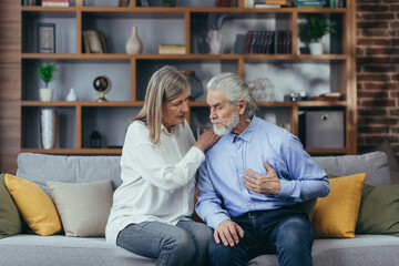 Senior mature woman helping her husband with chest pain. Old man feeling bad pain disease. Wife supporting husband - Elderly couple suffering from symptoms heart attack ache. sitting at home indoors