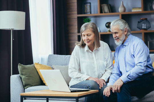 Senior Couple Man And Woman Shopping Online Browsing Laptop In Home. Elderly Family Happy Retired Couple Using Computerooking At Screen And Paying On Internet. Older Husband And Wife Choose The Web