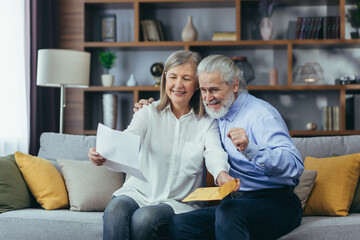 Happy senior retired couple sitting on sofa at home reading letter, happy and smiling