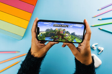 Fototapeta premium Girl holding a smartphone with Minecraft mobile game app on the screen. Blue background with school supplies, AirPods, video game controller. Rio de Janeiro, RJ, Brazil. April 2022