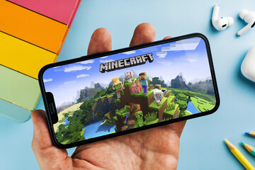Fototapeta premium Man holding a smartphone with Minecraft mobile game app on the screen. Blue background with school supplies, AirPods, video game controller. Rio de Janeiro, RJ, Brazil. April 2022