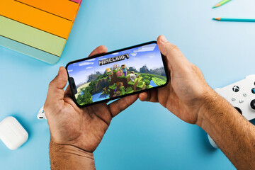 Fototapeta premium Man holding a smartphone with Minecraft mobile game app on the screen. Blue background with school supplies, AirPods, video game controller. Rio de Janeiro, RJ, Brazil. April 2022