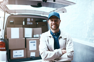 Fototapeta premium All packed up and ready for delivery. Portrait of a cheerful young delivery man standing next to a van full of boxes with his arms folded outside during the day.