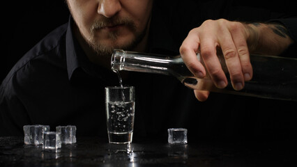 Alcoholic guy pouring up frozen vodka from bottle into shot glass with ice cubes on black background. Man drinking cold transparent alcohol drink sake, tequila or rum. Alcohol dependence addiction