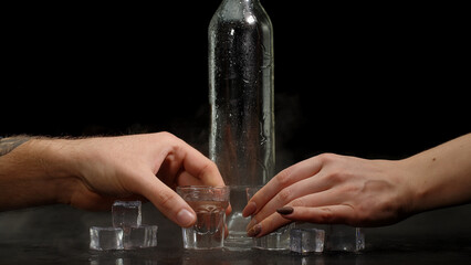 Bartender pouring up two shots of vodka with ice cubes from a bottle into glasses against black background. Two hands with glasses of vodka making cheers. Raising toast with glasses of tequila or sake