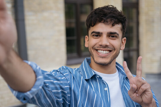 Handsome  smiling Indian man holding mobile phone taking selfie showing victory sign on the street. Happy stylish asian influencer communication online with subscribers outdoors