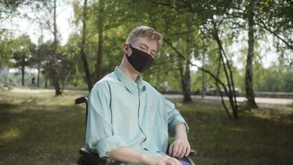 Being in the public park, a disabled white man in a blue shirt and a protective mask is sitting a wheelchair with a thumbs up and looking at the camera