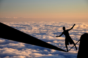 Silhouette of young man balancing on slackline high above clouds and mountains. Slackliner balancing on tightrope during sunset, highline silhouette.