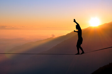 Silhouette of young man balancing on slackline high above clouds and mountains. Slackliner balancing on tightrope during sunset, highline silhouette.