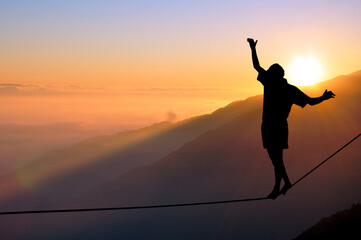 Silhouette of young man balancing on slackline high above clouds and mountains. Slackliner...
