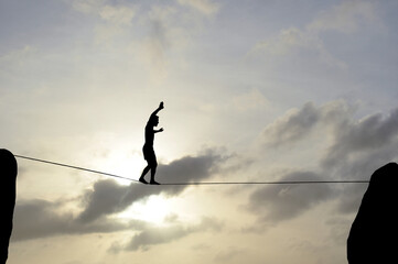 Silhouette of young man balancing on slackline, sun and clouds behind. Slackliner balancing on tightrope between two rocks, highline silhouette.