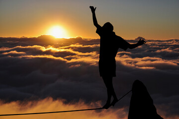 Silhouette of young man balancing on slackline high above clouds and mountains. Slackliner balancing on tightrope during sunset, highline silhouette.