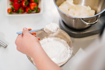 top view. A cook adds a spoonful of flour to prepare dough in a mixer bowl.