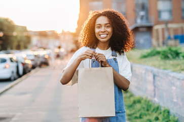  Takeaway food concept. Smiling african woman holds craft paper bag outdoors. Pick up food from a...