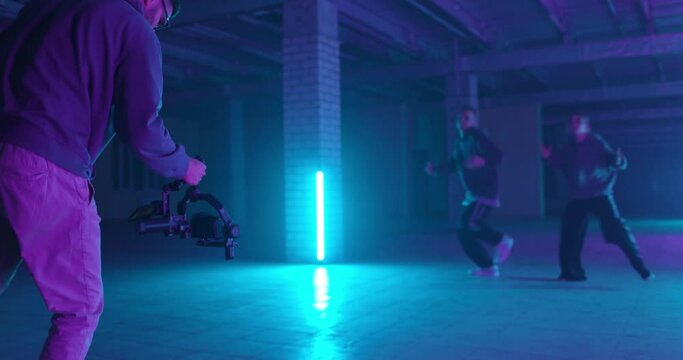 Backstage of video shooting. Cameraman filming hip-hop dancers in neon light. Operator moves with image stabilizer in hands at dark studio. Shooting a clip