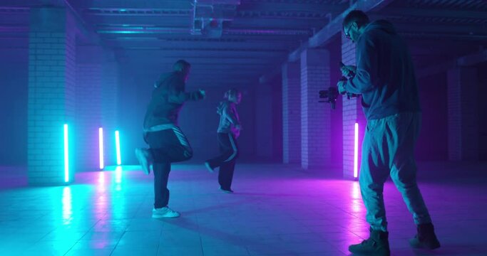 Backstage of video shooting. Cameraman filming hip-hop dancers in neon light. Operator moves with image stabilizer in hands at dark studio. Shooting a clip. Slow motion