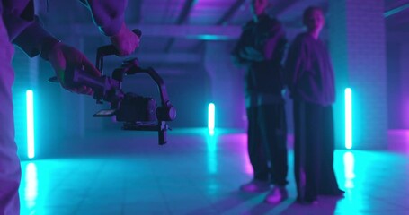 Backstage of video shooting. Cameraman filming hip-hop dancers in neon light. Operator moves with image stabilizer in hands at dark studio. Shooting a clip. Back view
