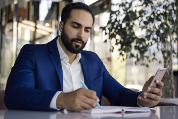 Pensive handsome Arabian businessman holding mobile phone taking notes working in office. Busy middle easter manager planning startup sitting at workplace. Successful business 