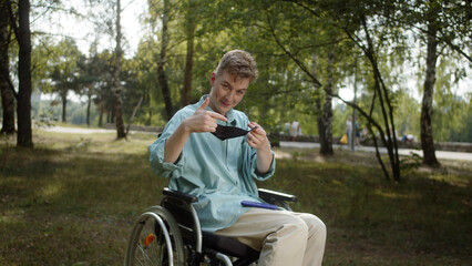 Fototapeta premium A disabled white man iin a blue shirt s sitting in a wheelchair and putting a protective mask on his dace, slow motion