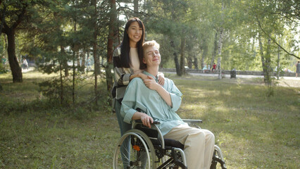 Fototapeta na wymiar A pretty asian lady with long dark hair hugs her disabled young man in a blue shirt, they look at the camera and smile