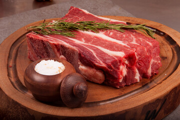raw meat cut with fat denver steak in butcher shop on wooden board with rosemary and coarse salt, anlge view, close-up